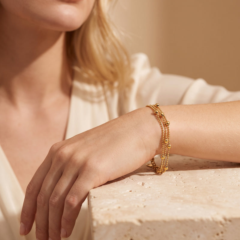 Gold-tone three-strand bead bracelet worn on a wrist over a textured light-colored block.