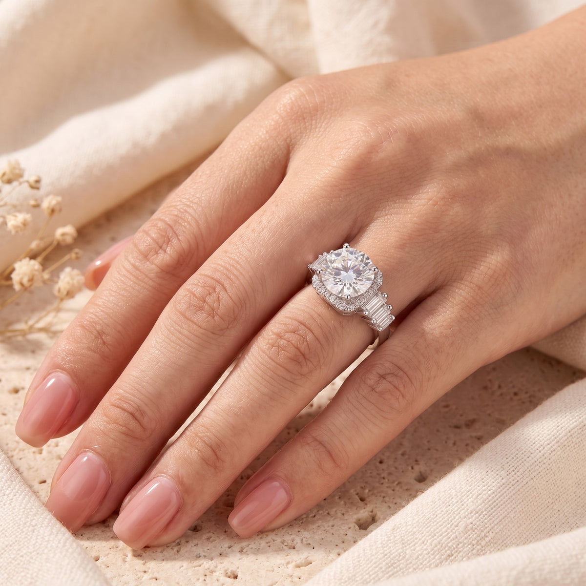 Silver cushion halo ring with baguette side stones shown on a hand against soft cream fabric.