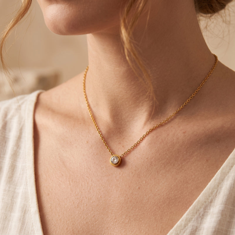 Woman wearing a gold-tone bezel solitaire necklace with a clear round center stone and a white top.