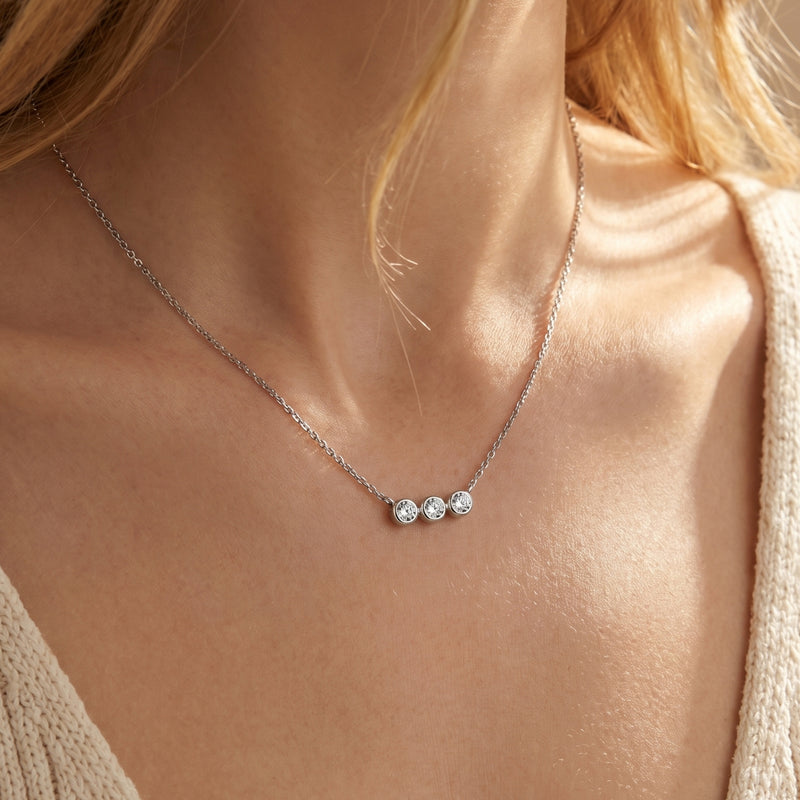 Silver three-stone moissanite bezel necklace worn by a model in a neutral sweater.