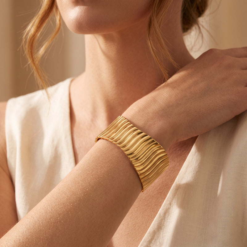 Gold-tone rippled cuff bracelet worn on a model’s wrist against a cream outfit.