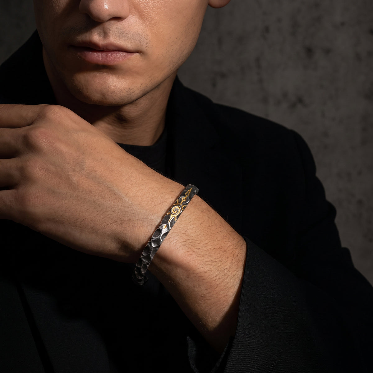 Man wearing the hammered silver cuff bracelet with a gold-tone sigil near his face, dressed in black.