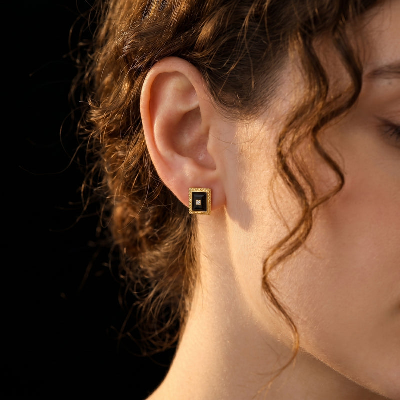 Woman wearing a yellow gold square-cut black onyx stud earring with a center accent stone.