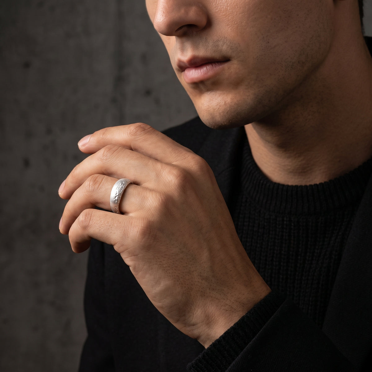 Model wearing a textured silver open ring on his ring finger with his hand raised near his chin.