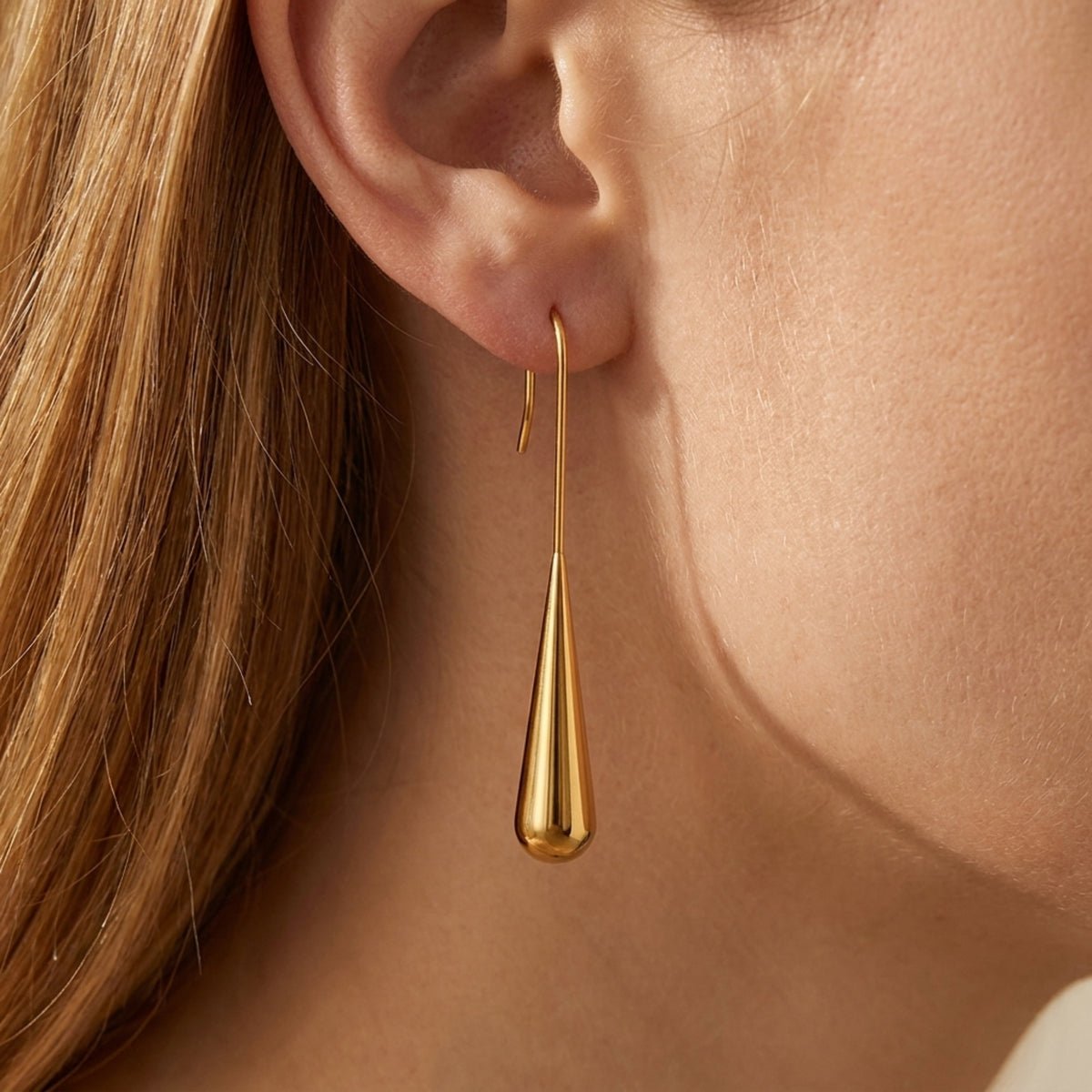 Close-up of a gold-toned teardrop earring hanging from a model's ear by a curved hook.