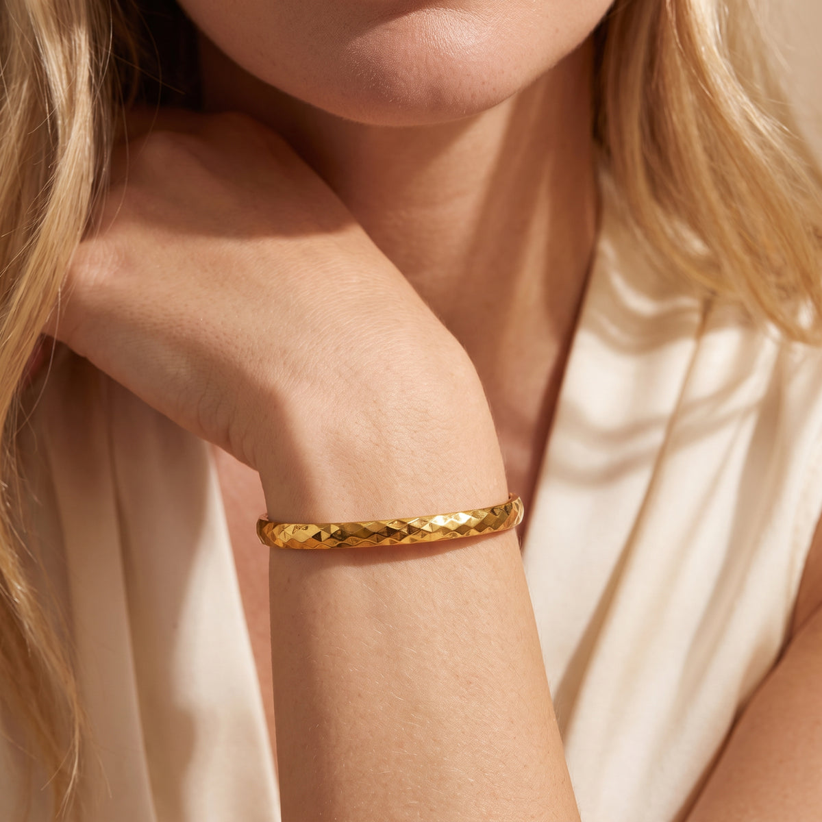 Gold-toned faceted bangle bracelet worn on a wrist beside the wearer's face, with light clothing in the background.