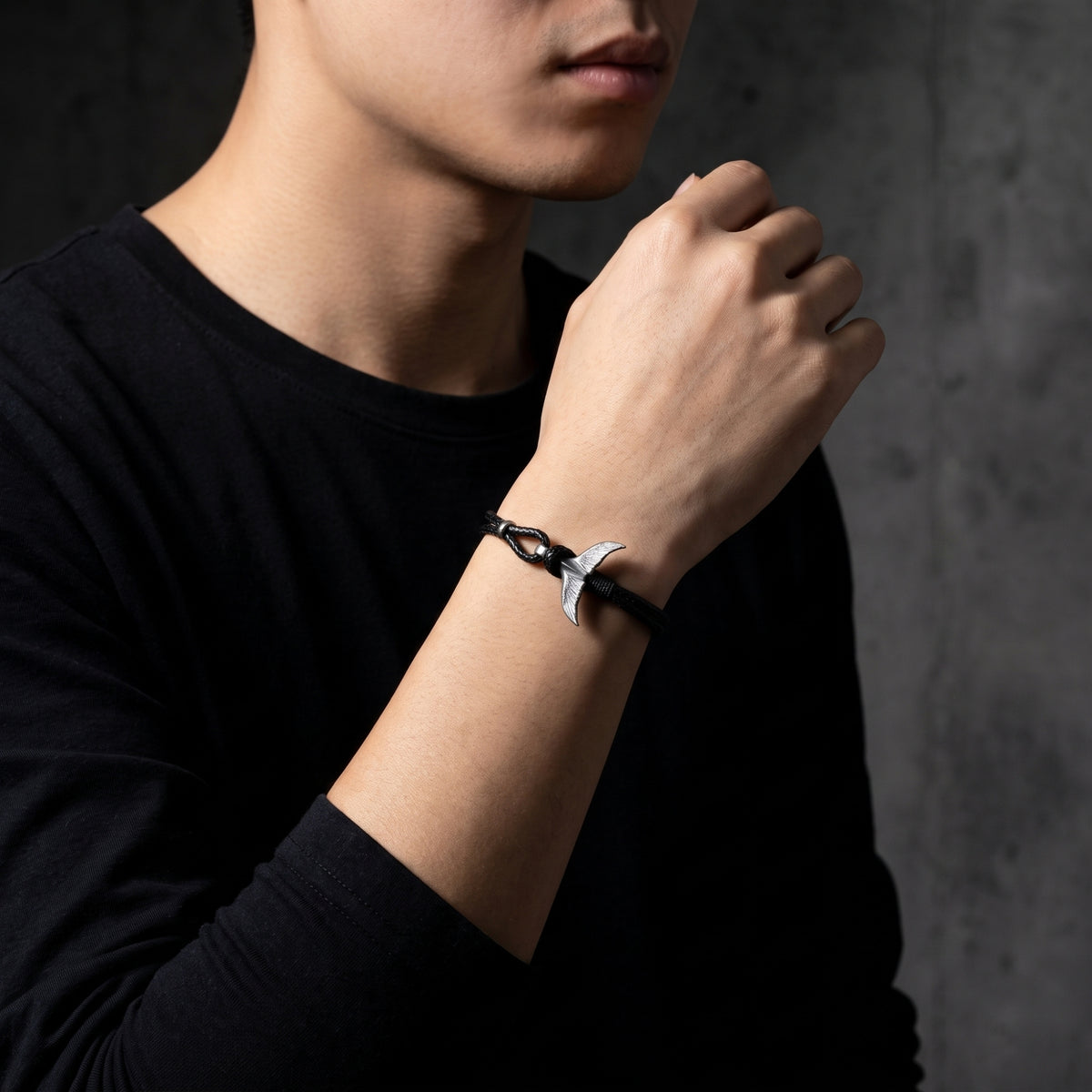 Model wearing the black cord bracelet with a silver whale tail clasp over a black long-sleeve shirt.