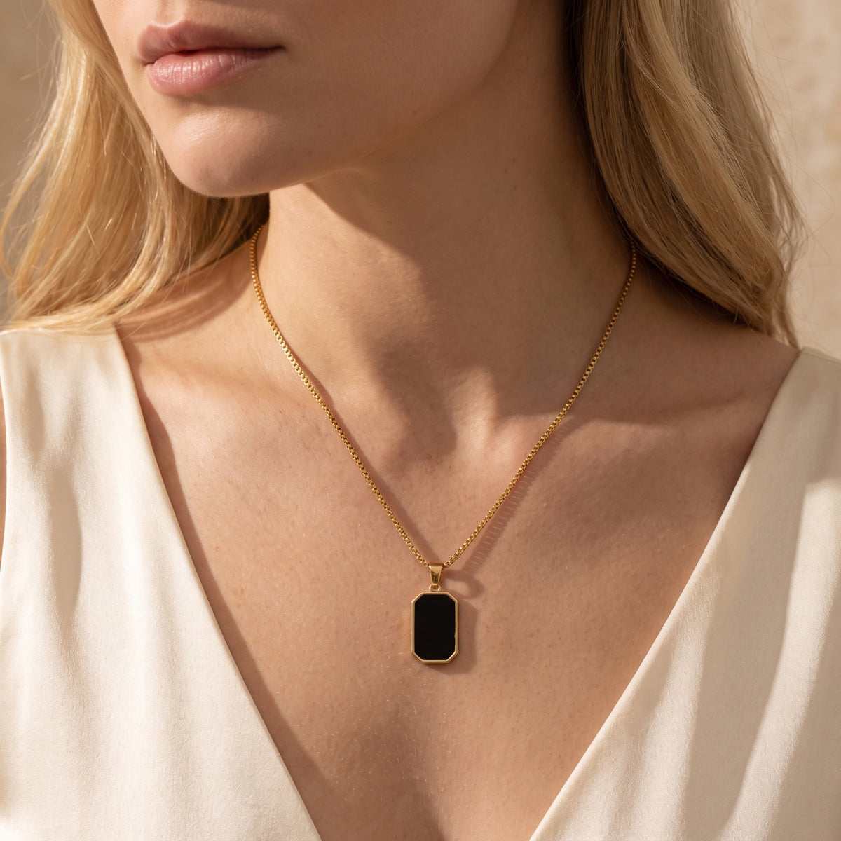 Model wearing a gold-tone box chain necklace with an octagonal black pendant over a cream V-neck blouse.