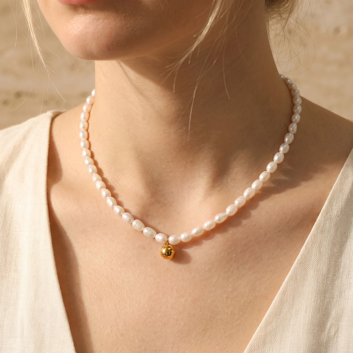 Model wearing a white pearl strand necklace with a dangling gold-tone sphere pendant on the chest.