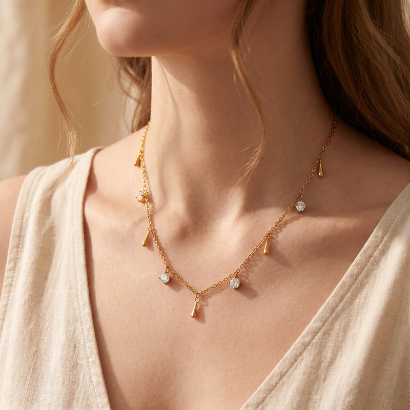 Gold-tone station necklace with clear stones and smooth droplet charms worn on a woman's neck.