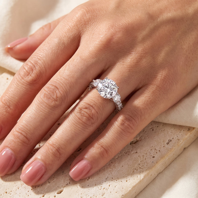 On-hand view of the silver three-stone ring with a round center stone, worn on the ring finger.