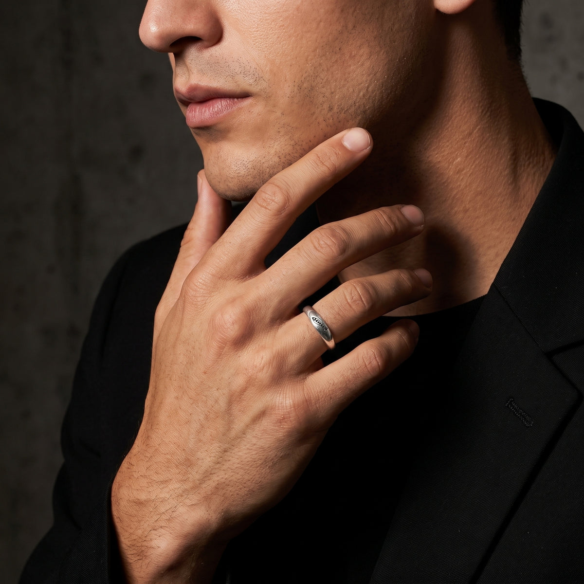Man wearing a silver carved fish ring while touching his jaw, dressed in a black blazer.