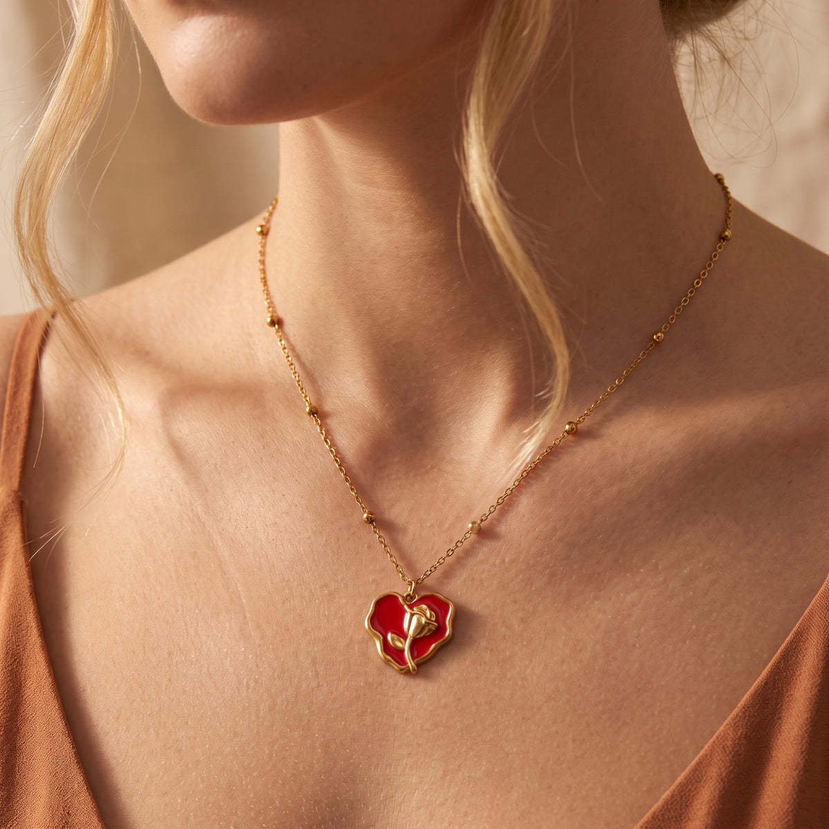 Model wearing the red enamel rose heart pendant necklace with a gold-tone beaded chain and rust-colored top.
