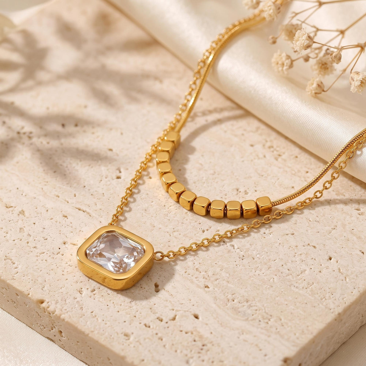Close-up of a gold-tone square clear pendant and cube-beaded chain resting on a stone slab.