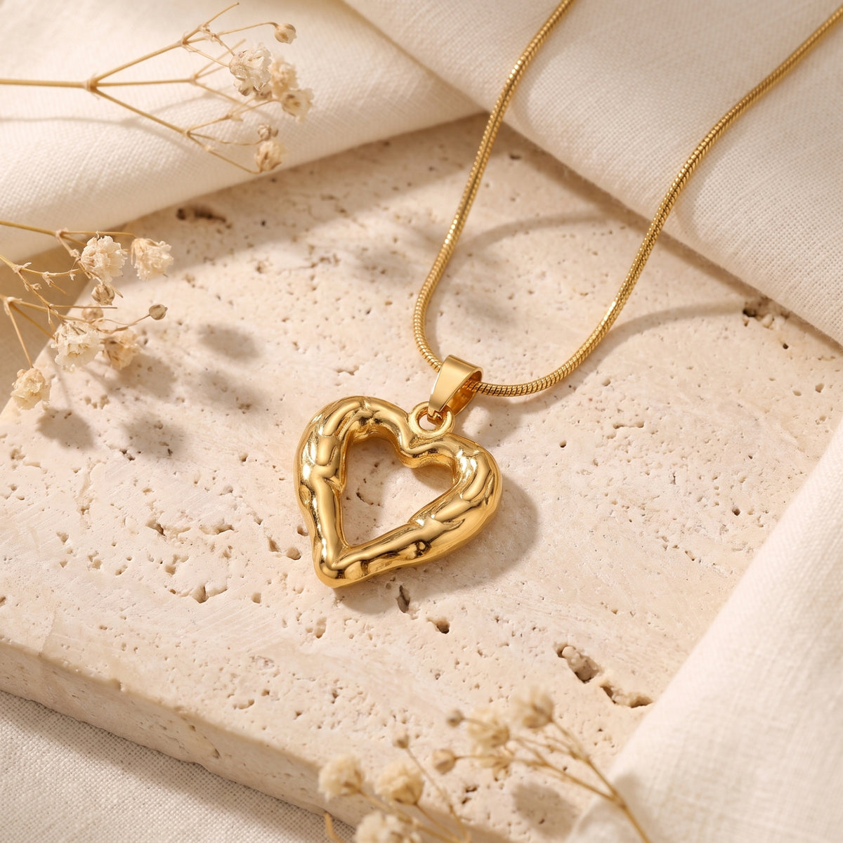 Gold-tone molten heart necklace resting flat on a travertine tile near white fabric and flowers.