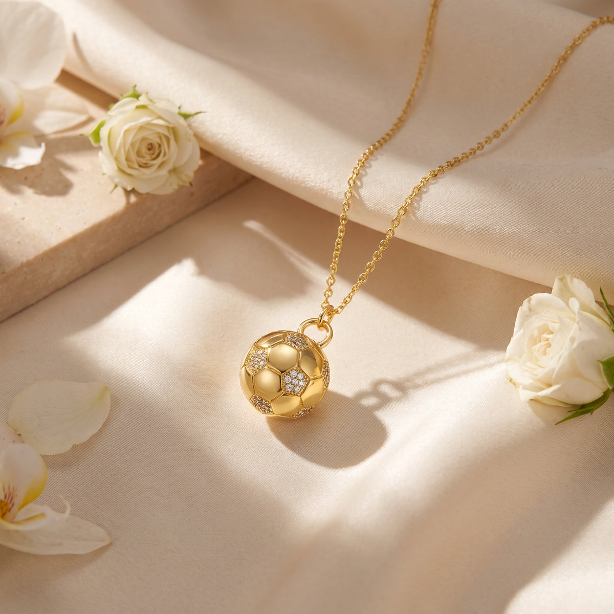 Gold-tone soccer ball necklace displayed on silky fabric surrounded by white rose blooms and petals.