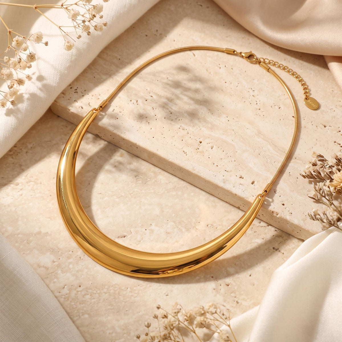 Flat lay of the complete gold-tone crescent collar necklace on a beige stone surface, showing its chain and clasp.