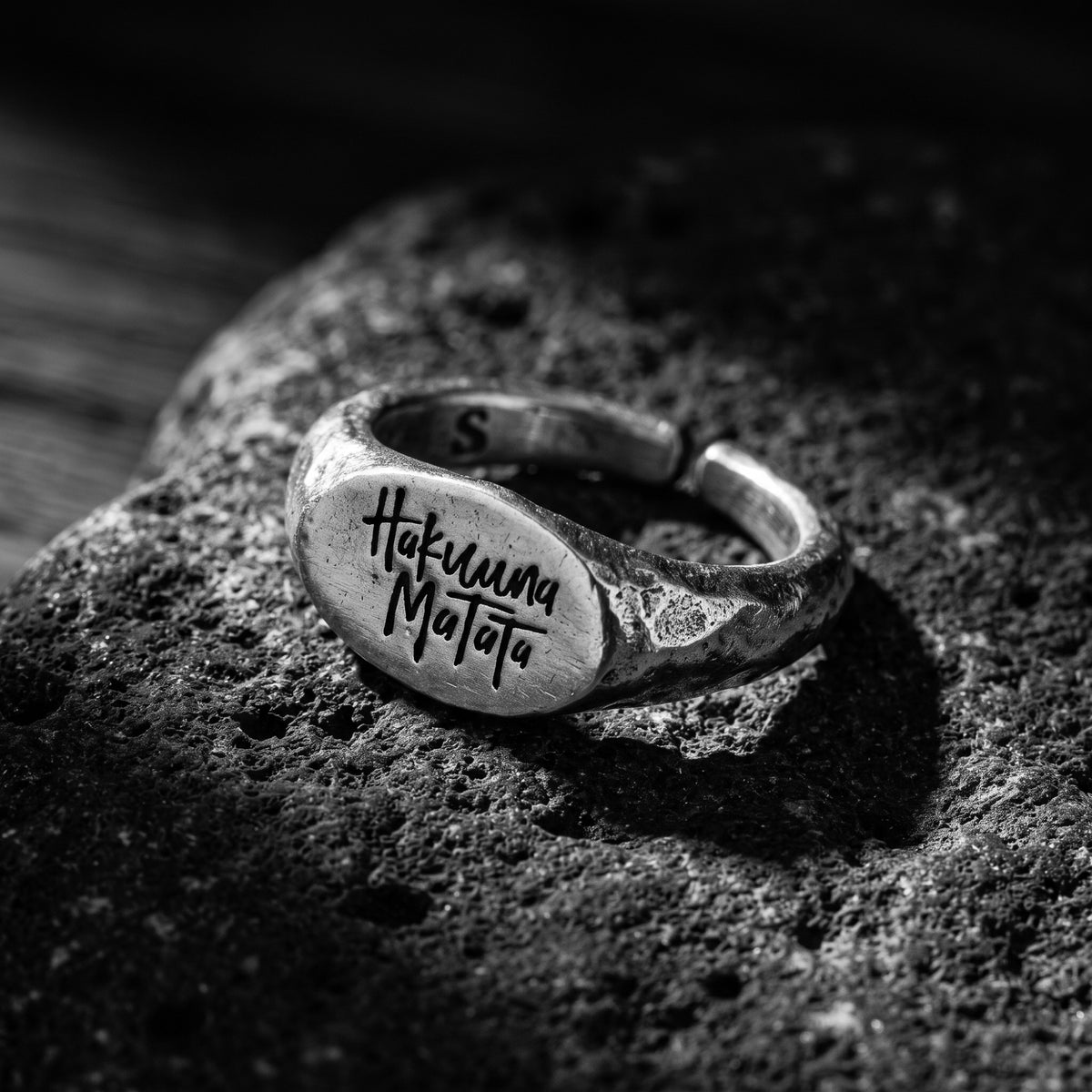 Angled view of the silver open-band Hakuna Matata signet ring resting on textured dark rock.