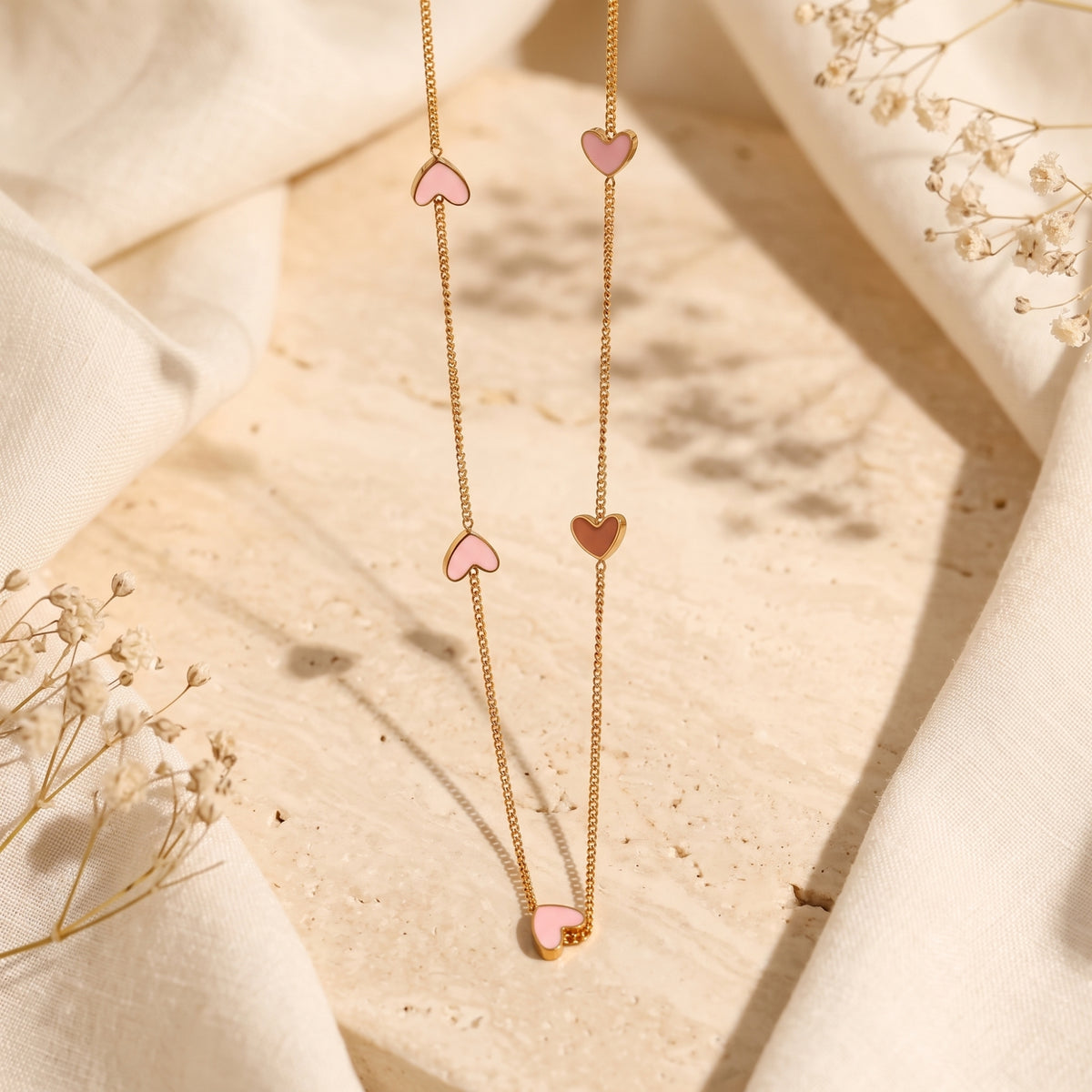 Gold-tone station necklace with pink and rose enamel heart stations laid vertically on stone and linen.