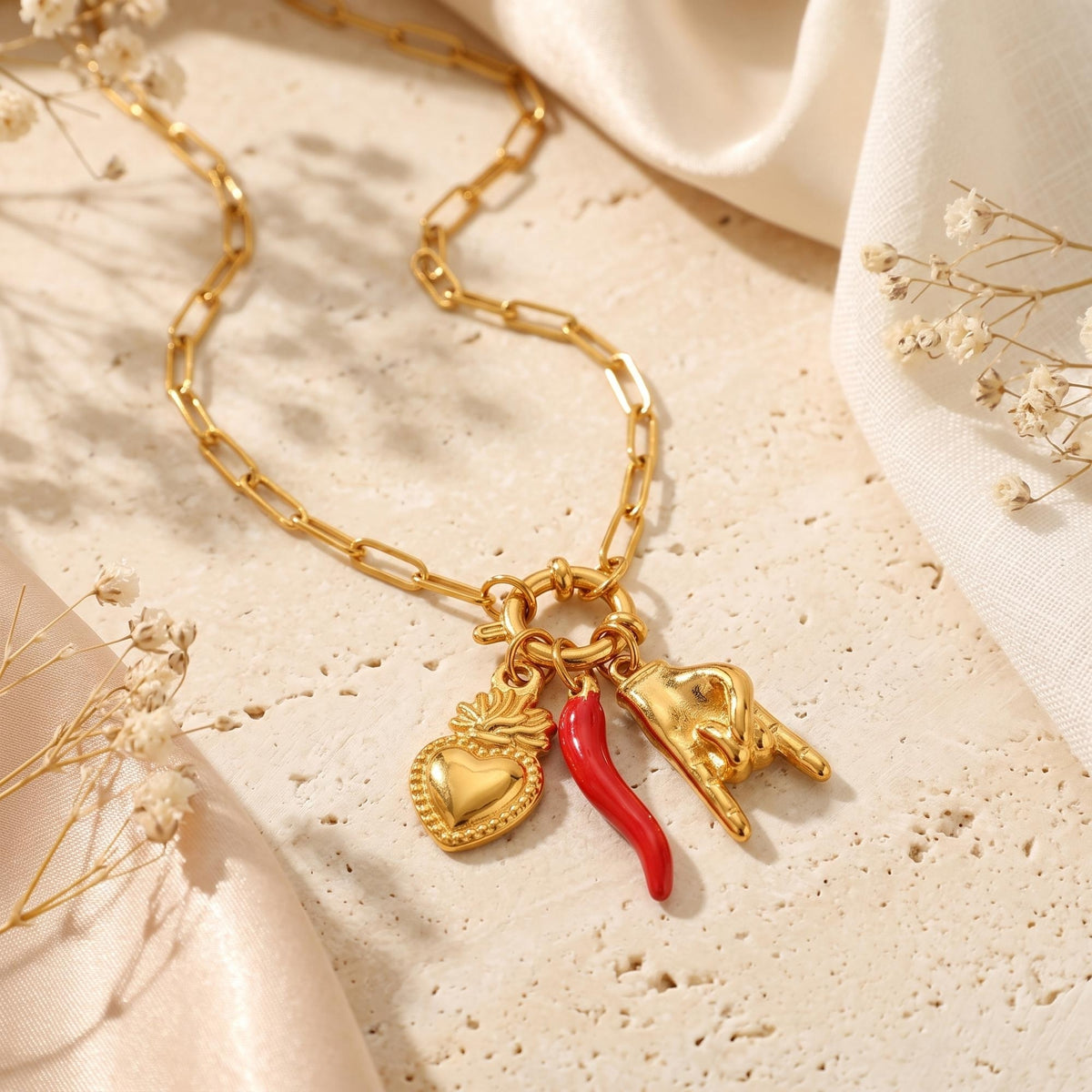 Flat lay of the gold-tone paperclip charm necklace with heart, red pepper, and hand charms on a beige stone surface.