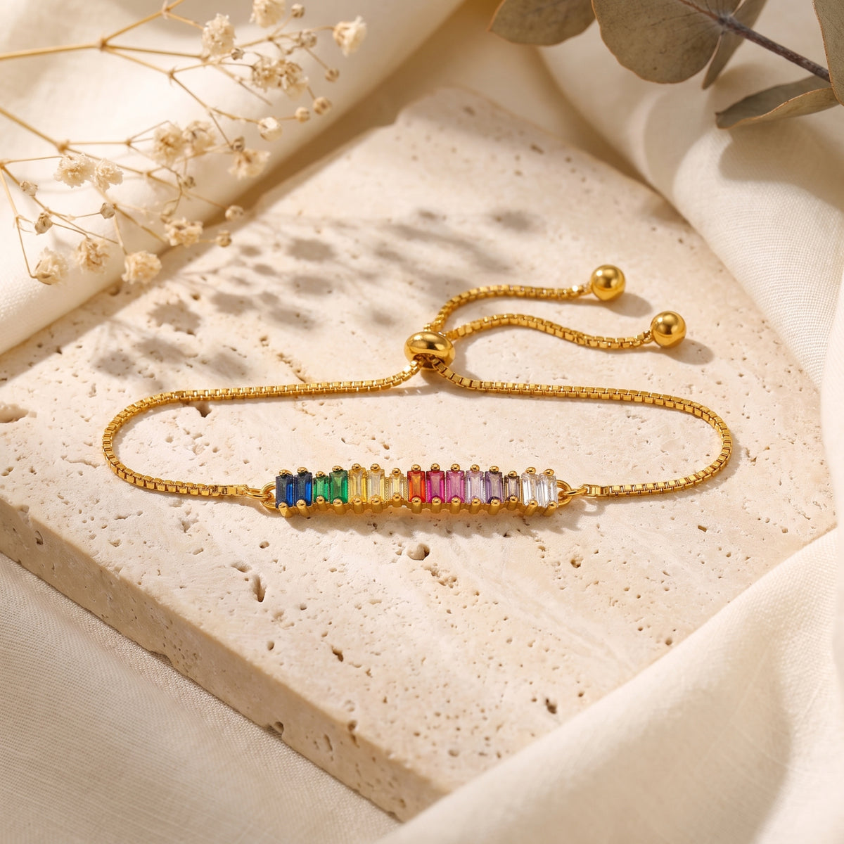 Full view of the gold-tone slider bracelet laid in a wide oval on stone, showing rainbow baguette stones.