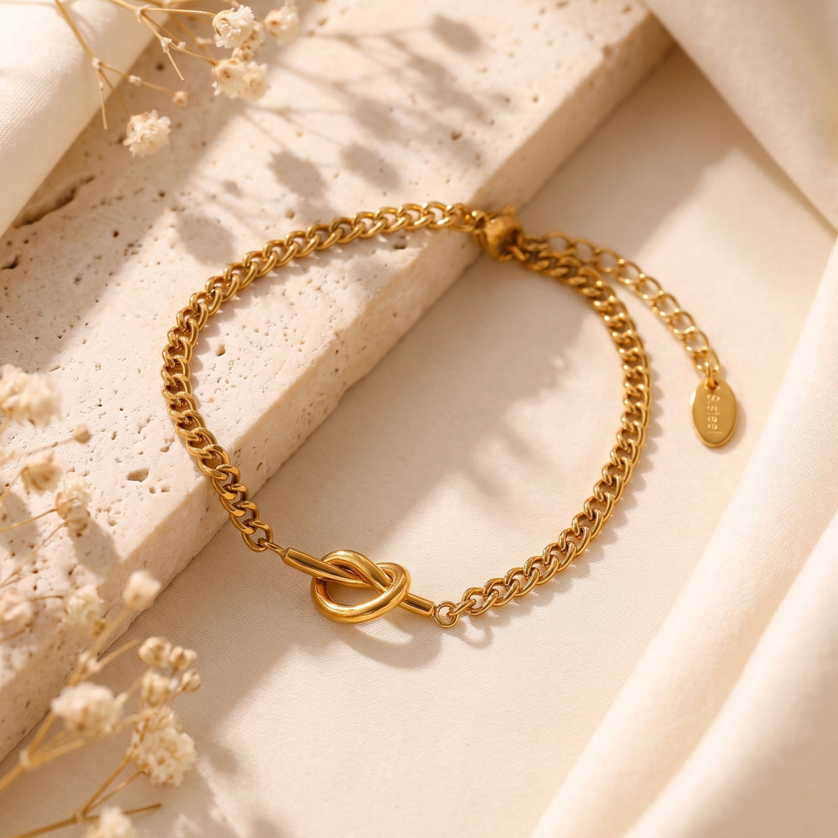 Gold-tone knot link bracelet arranged on a stone slab, with its extender chain and oval steel tag visible.