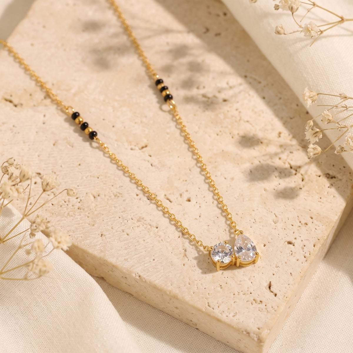Gold-tone chain necklace with round and pear-cut clear stones displayed flat on a textured beige stone block.