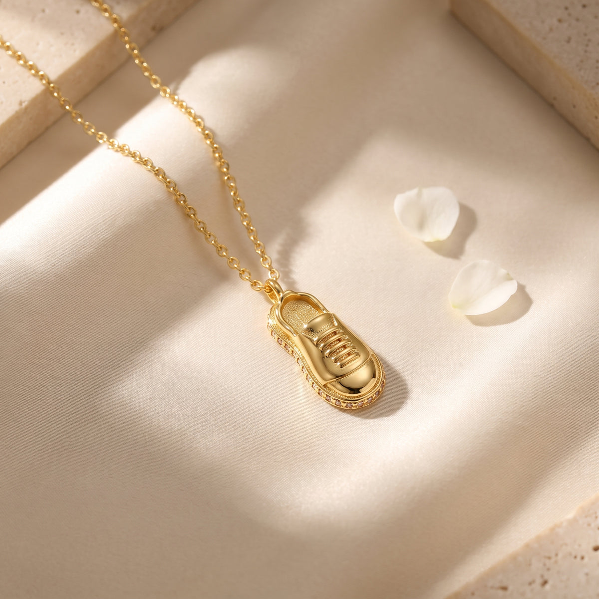 Gold sneaker pendant necklace lying flat on soft cream fabric beside white flower petals.