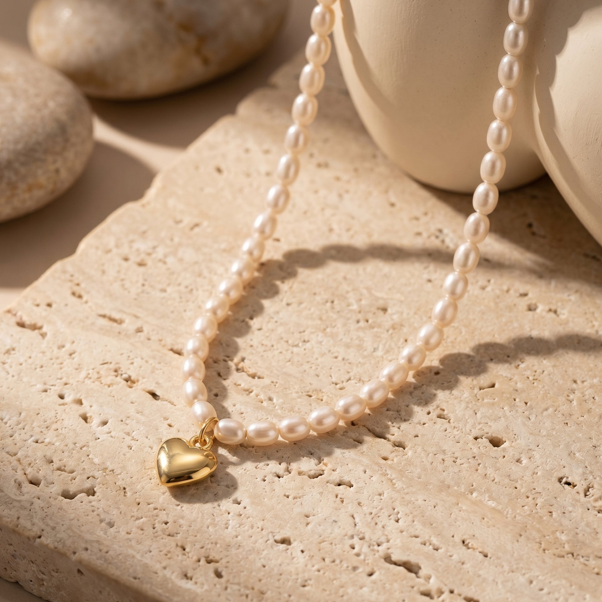 The Colette Pearl Necklace, a solid yellow gold heart charm and white synthetic pearl necklace, styled on a neutral stone background.
