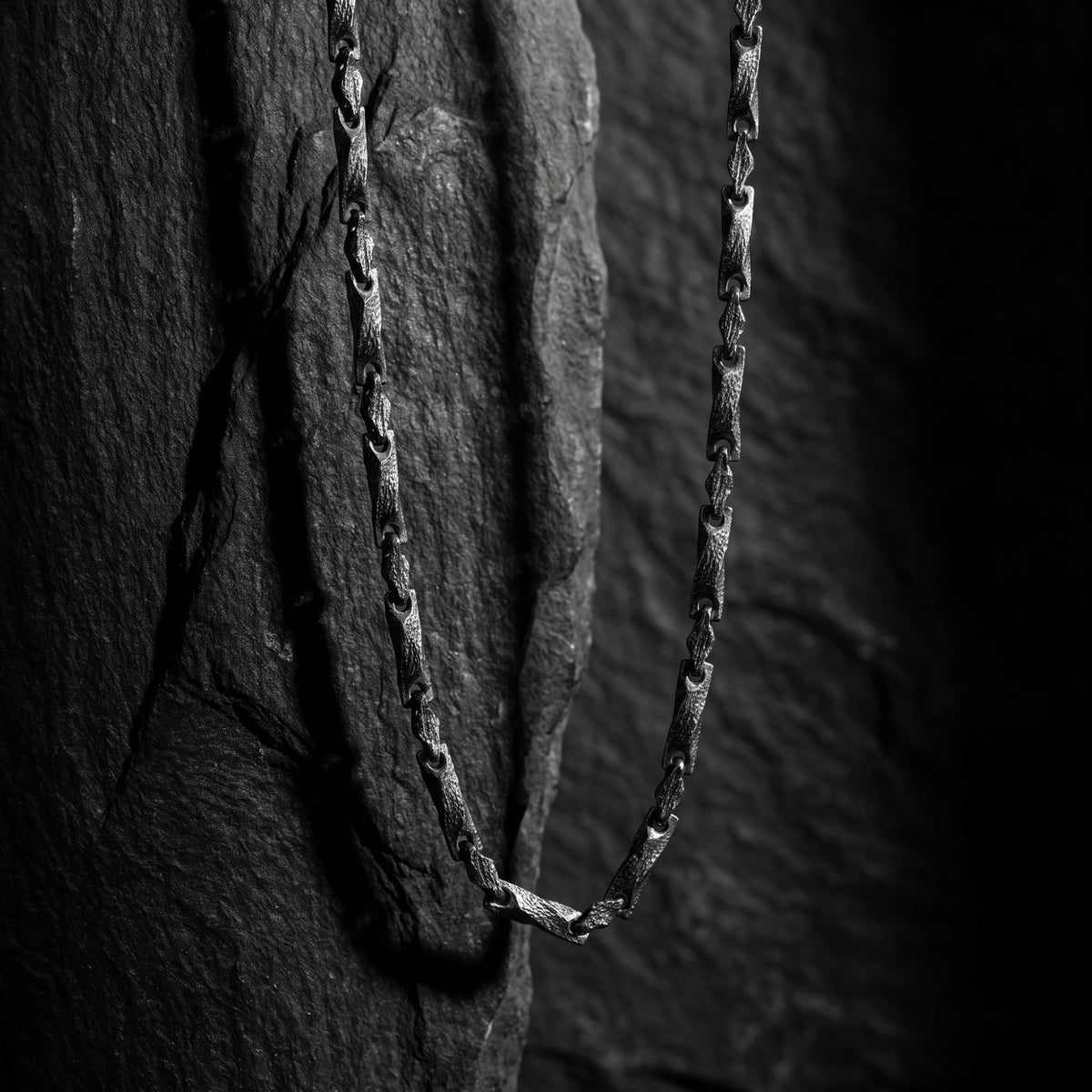 Textured silver bar link chain hanging vertically against a dark rock backdrop.