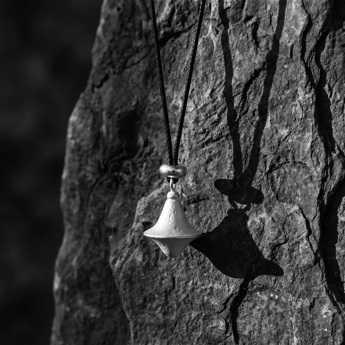 Black-and-white view of the textured silver double-cone pendant hanging against a rough stone surface.