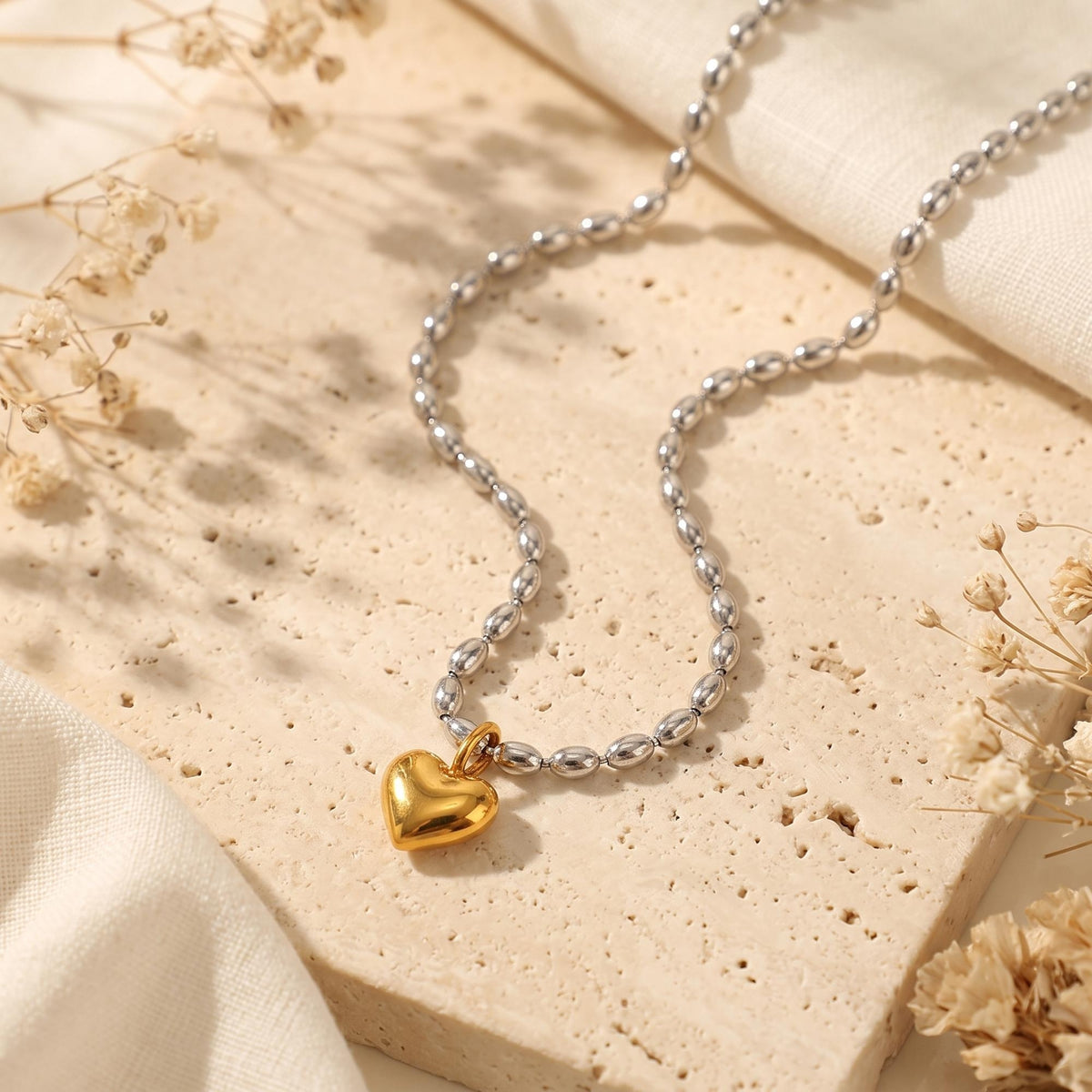 Full view of the two-tone puffed heart bead necklace curved on a textured beige slab with dried florals.