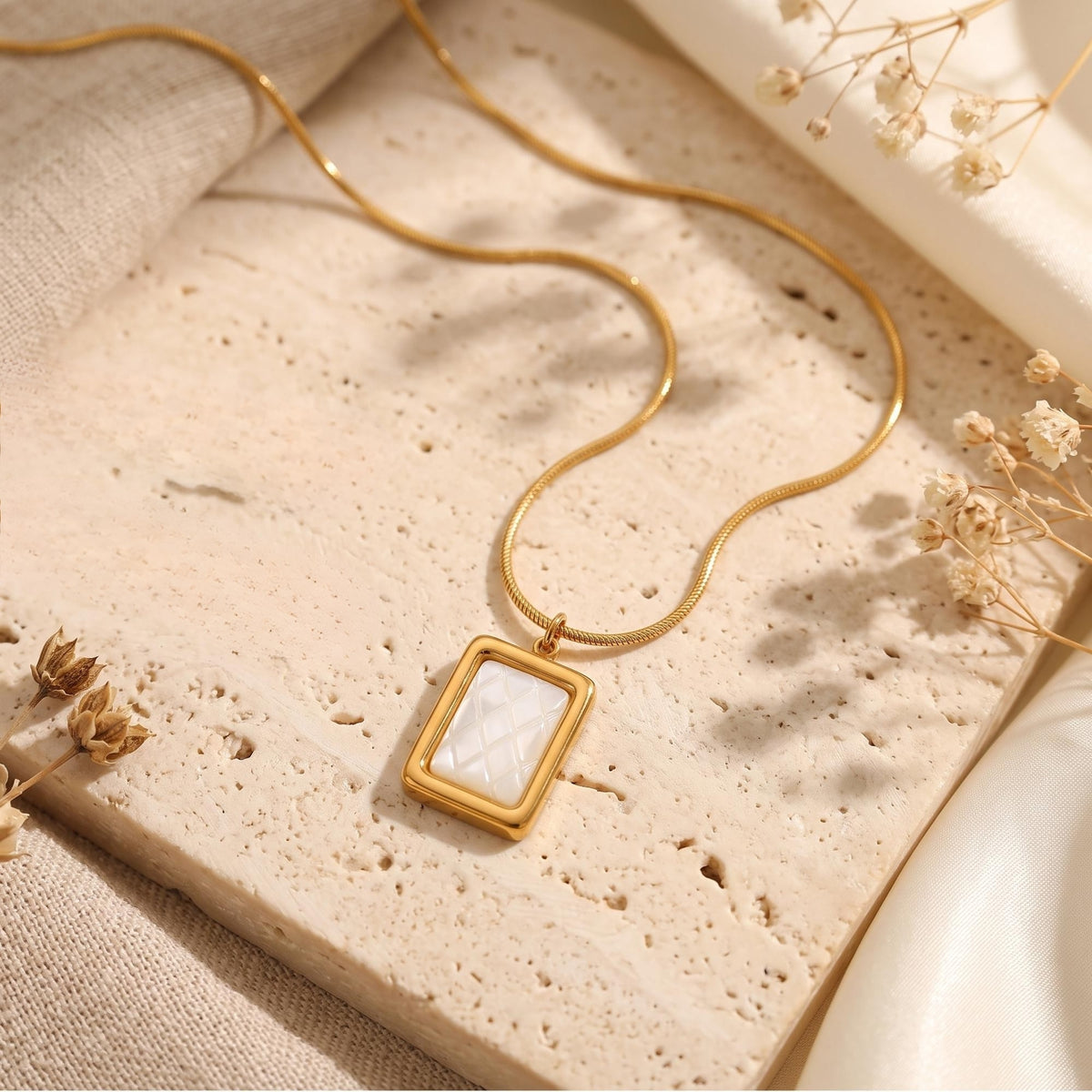 Close-up of a rectangular gold-tone pendant necklace with a quilted white inlay laid on a textured stone slab.