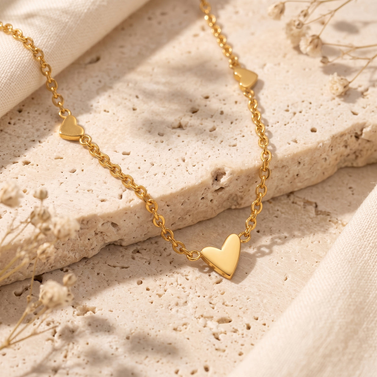 Close-up flat lay of the gold-tone heart station necklace on a beige stone slab with dried flowers.