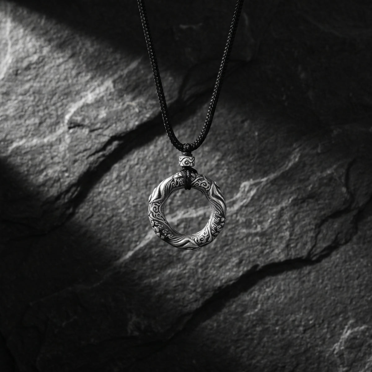 Front view of the carved circular silver pendant hanging on a black cord against a dark stone surface.