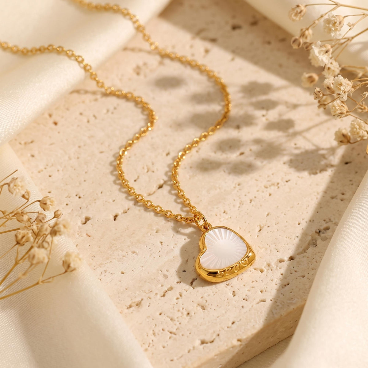 Gold-tone sunburst heart necklace laid flat on stone, surrounded by ivory silk and dried flowers.