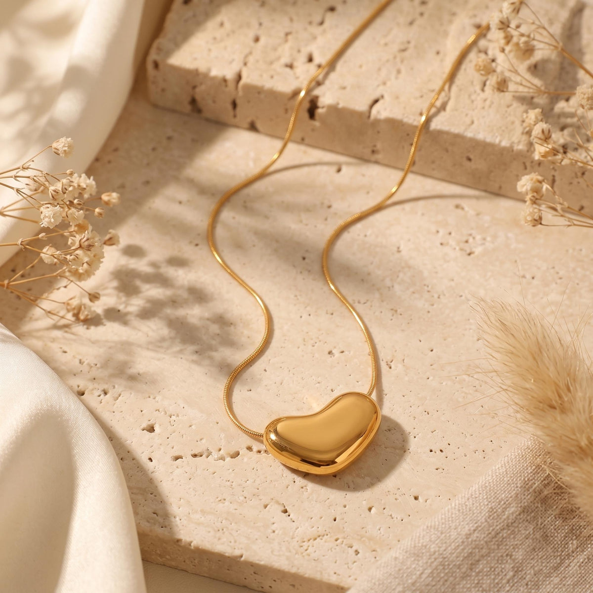 Gold-tone heart pendant necklace displayed flat with a curving snake chain on stone and cream fabric.