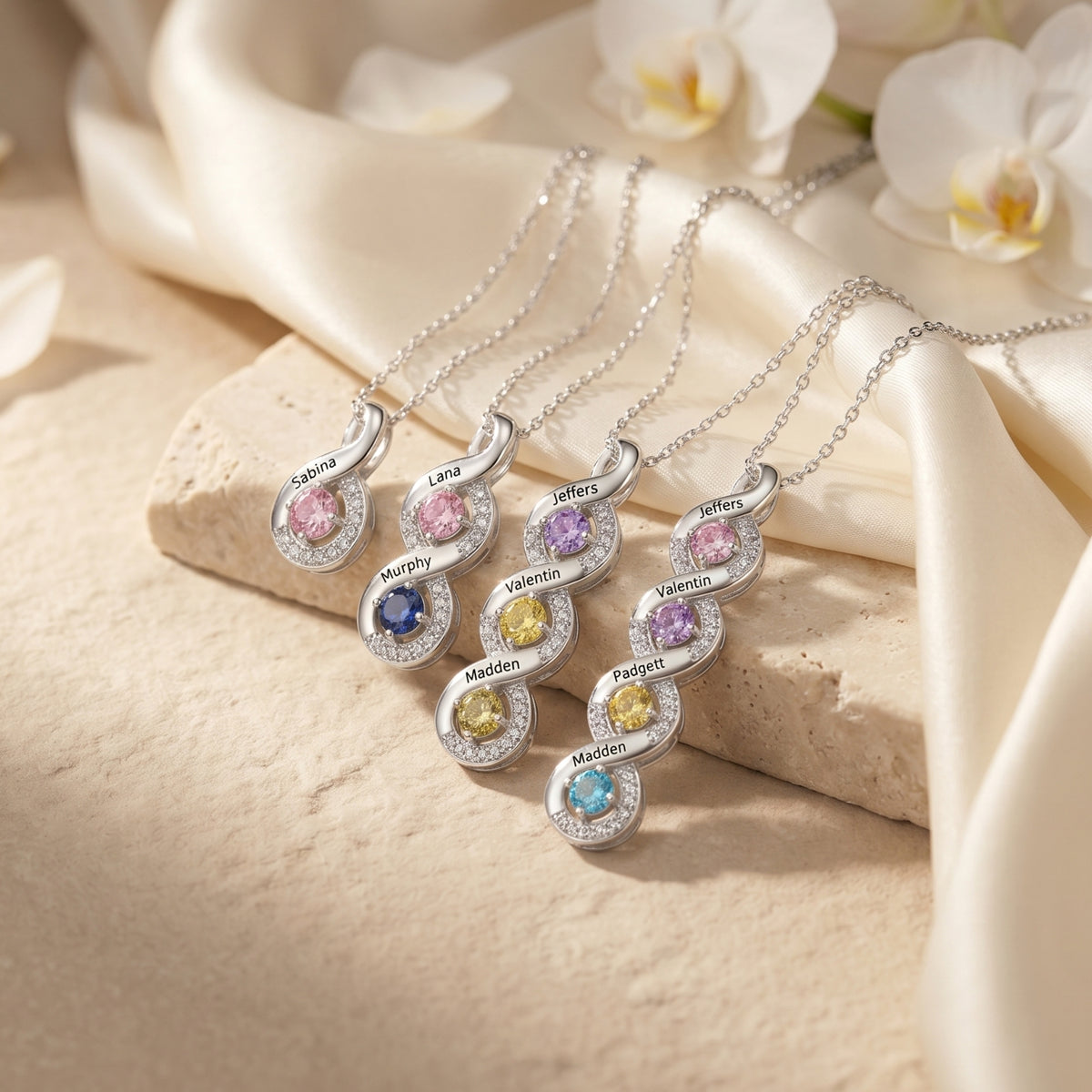 Four silver custom name infinity necklaces with various colorful birthstones on a textured surface.