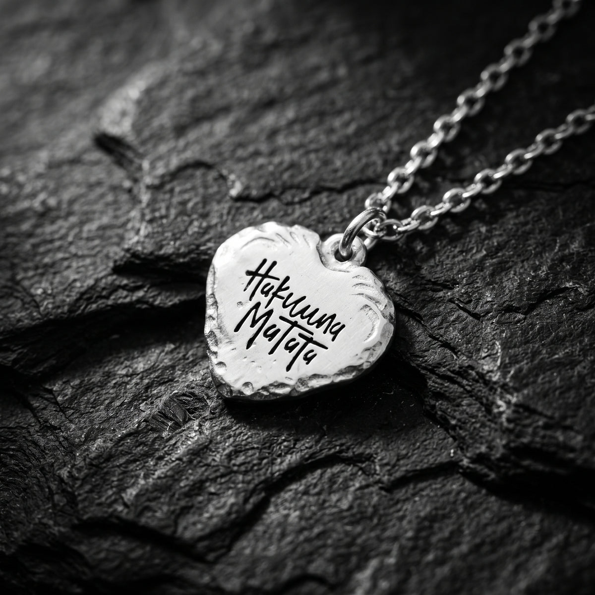 Angled close-up of the silver heart pendant resting on dark slate with the “Hakuna Matata” inscription visible.