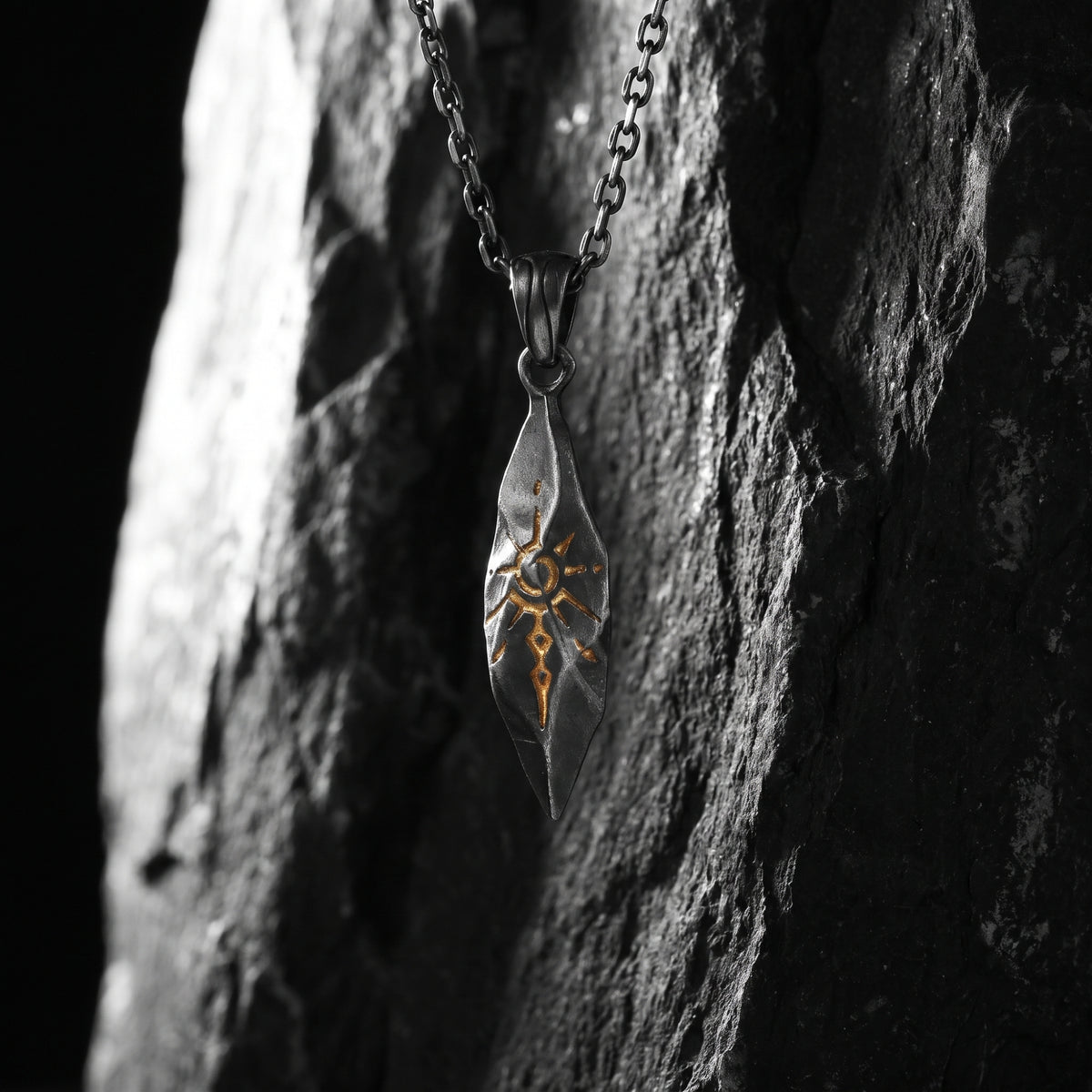 Silver talisman pendant hanging vertically against dark stone with a gold-colored sunburst motif