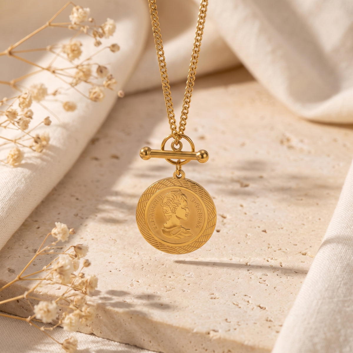 Gold-tone coin medallion necklace hanging above a pale, porous stone block with the T-bar clasp visible.