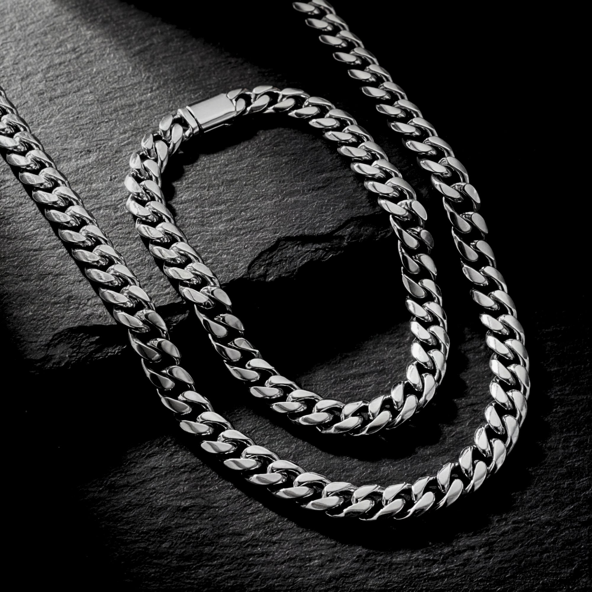Angled view of the silver stainless steel Cuban link chain and bracelet set on a dark stone surface.