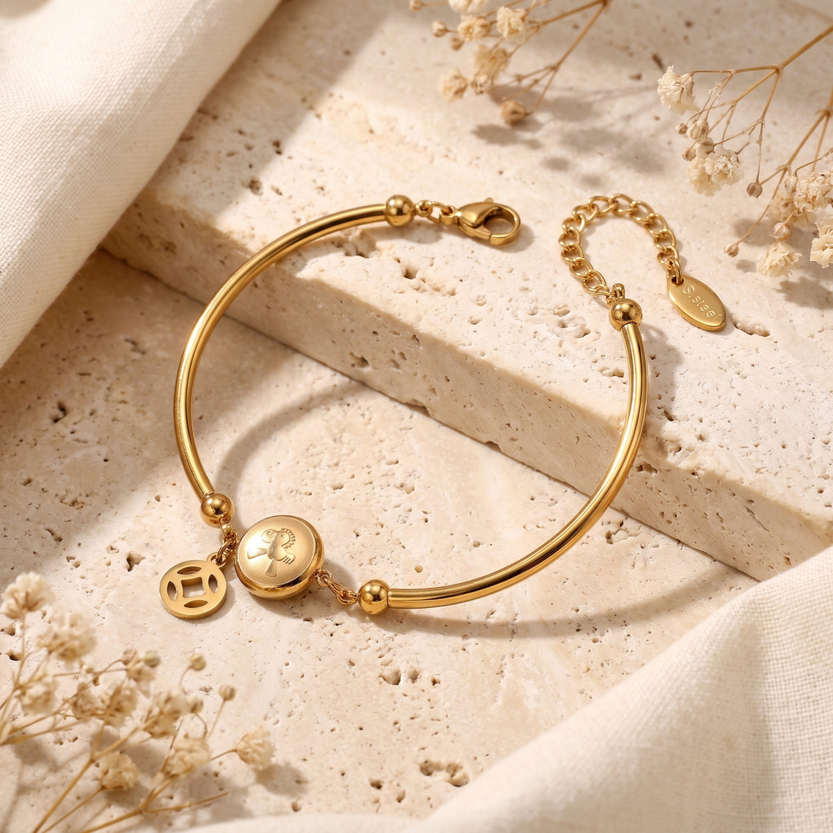 Gold-tone station bracelet displayed on a stone ledge showing the lobster clasp, extender chain, and oval S.Steel tag.