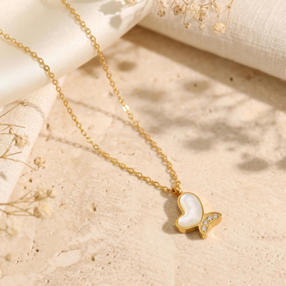 Gold butterfly necklace with mother of pearl inlay styled on a stone surface with dried flowers.
