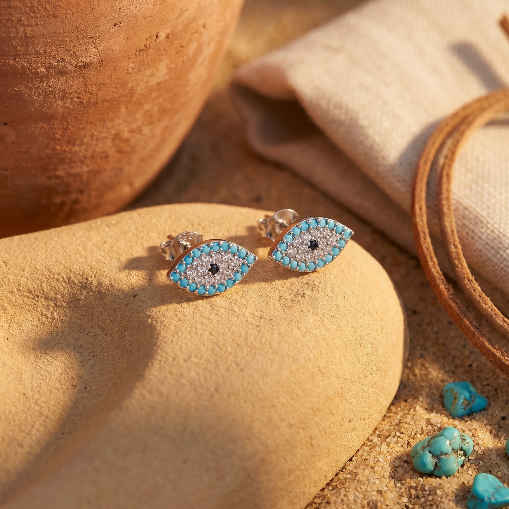 Pair of silver turquoise evil eye stud earrings displayed on a desert stone with sand and Southwestern pottery.