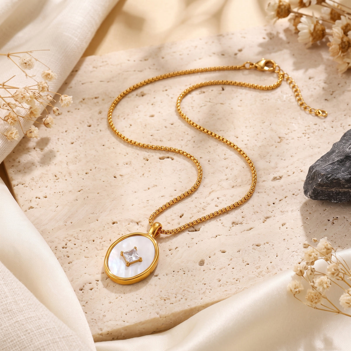 18K gold Lucelle necklace with mother of pearl pendant displayed on a stone slab with dried flowers and cream fabric.