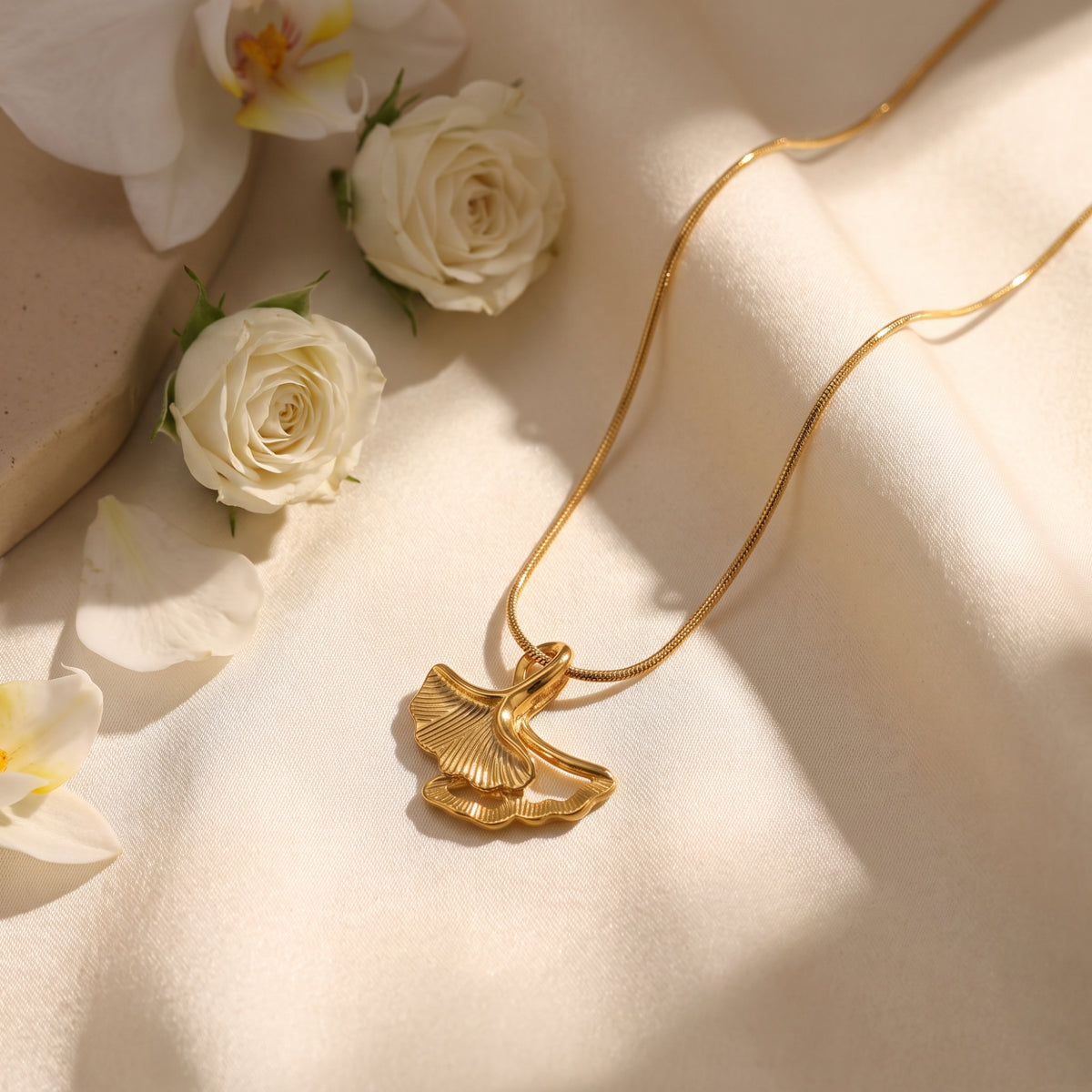 Close-up of the gold-tone double ginkgo leaf pendant on draped ivory fabric beside white flowers.