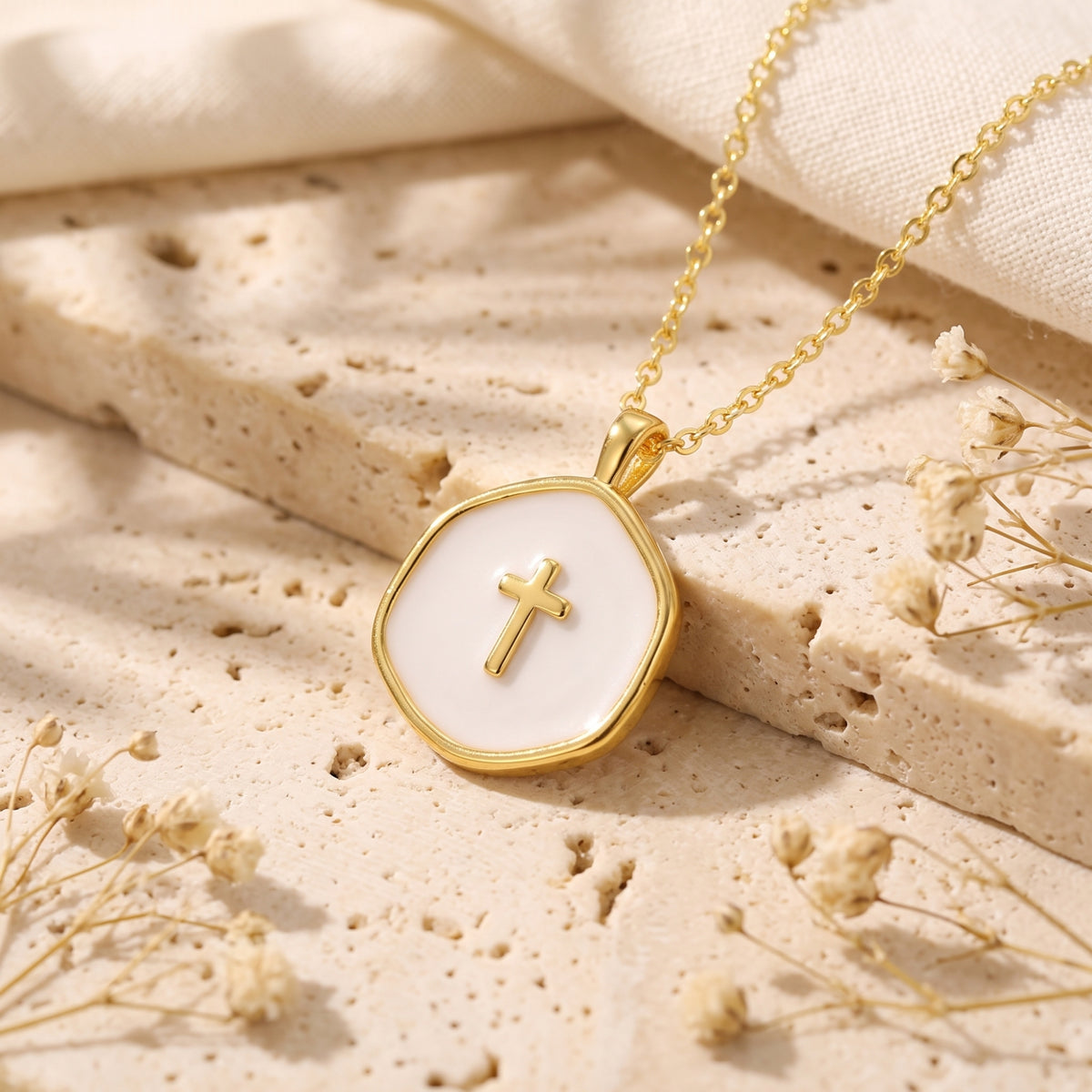 Angled close-up of a gold-tone cross pendant with a white enamel center on a light stone surface.