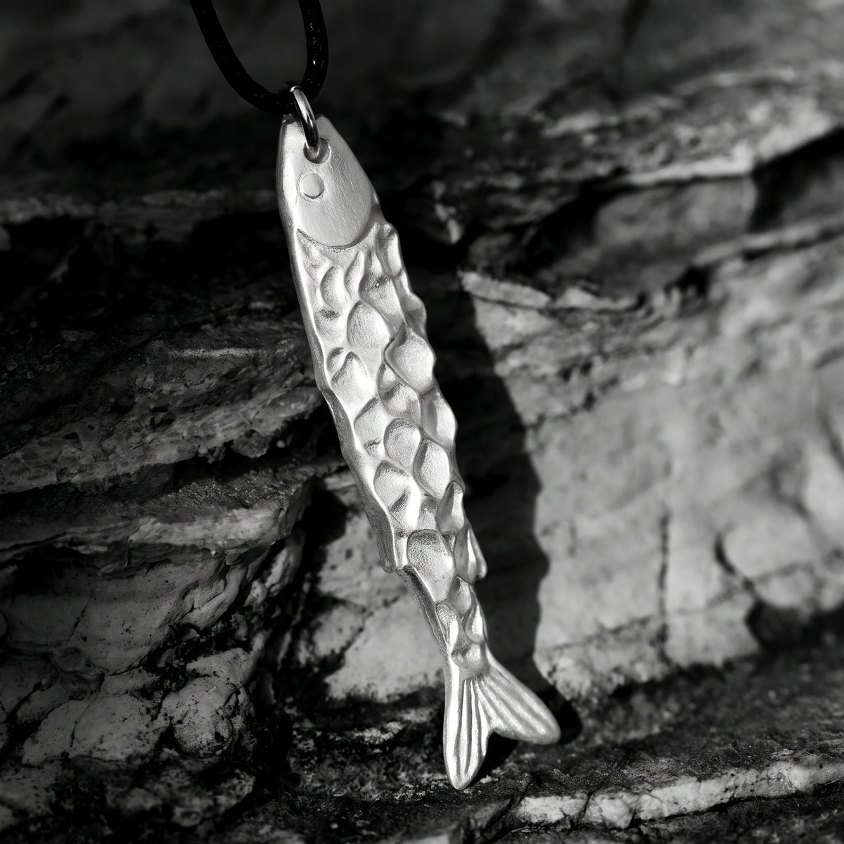 Close-up of the sculpted silver fish pendant hanging vertically against a rough grey stone background.