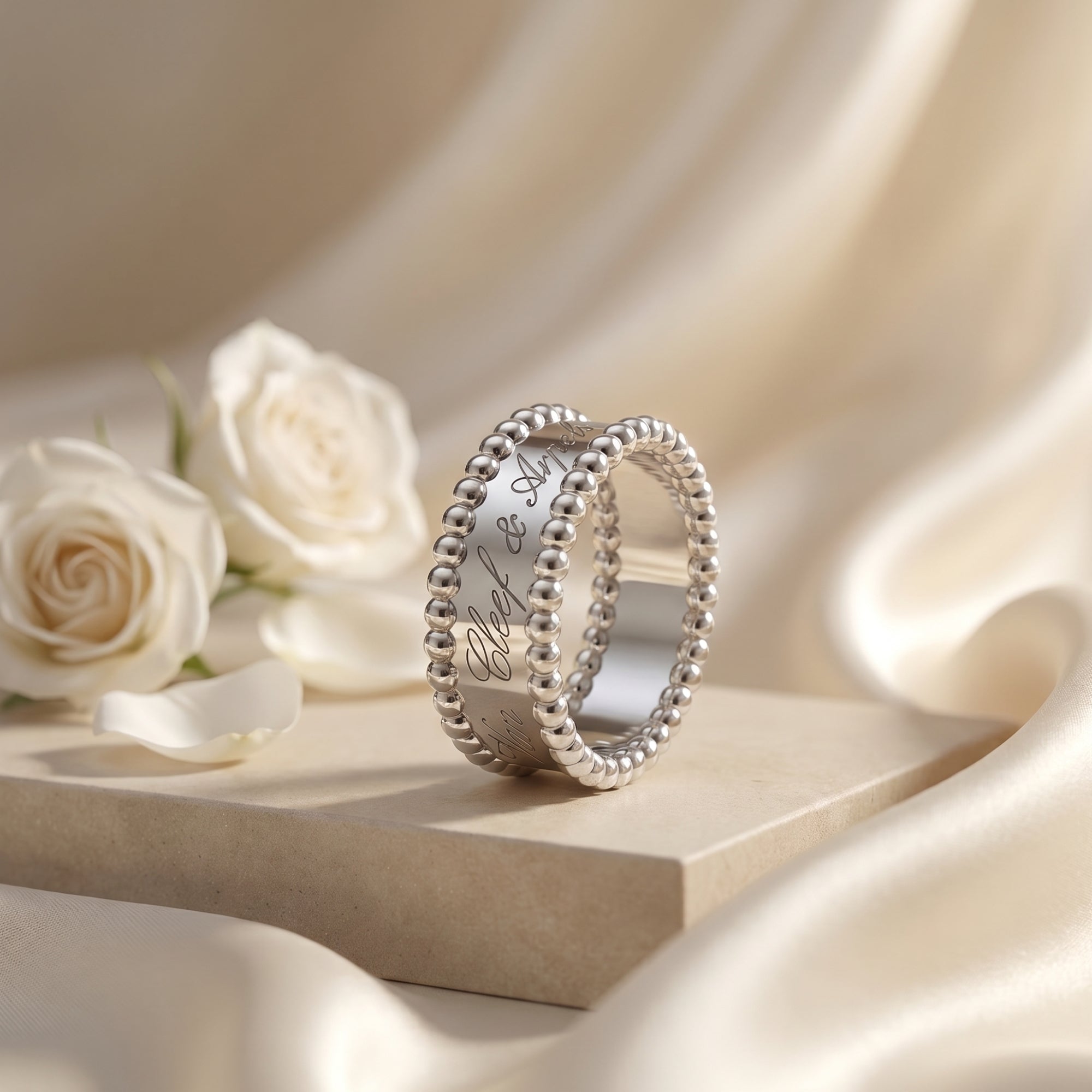 The Vowbound Ring with a silver finish and beaded edges displayed next to white roses on a silk background.